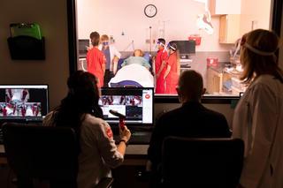 Three clinicians observe diagnostic screens and students in a simulated hospital room from behind glass. On the other side of the observation glass, students tend to a manikin.